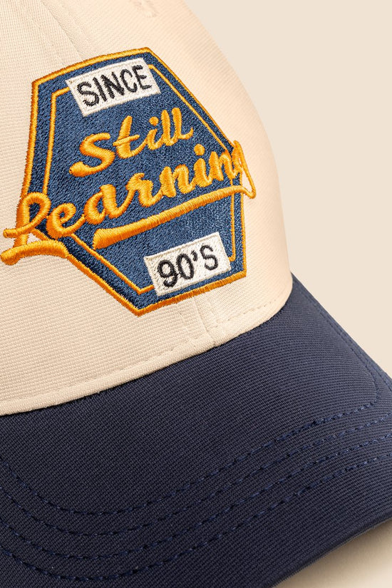 Cap with 'Since Still Learning 90s' embroidered patch on beige and navy background