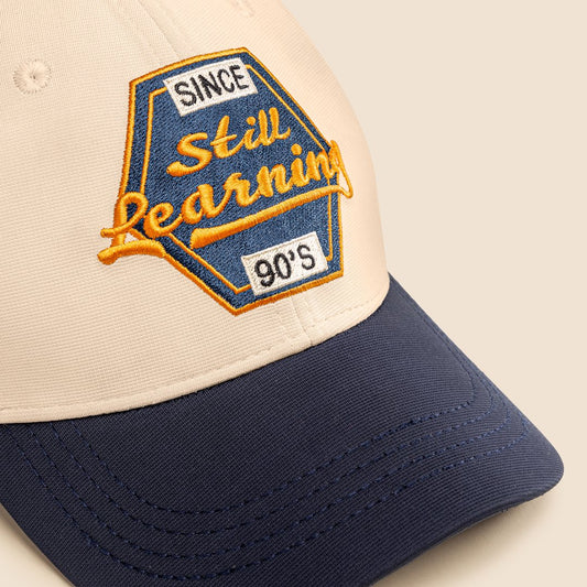 Cap with 'Since Still Learning 90s' embroidered patch on beige and navy background